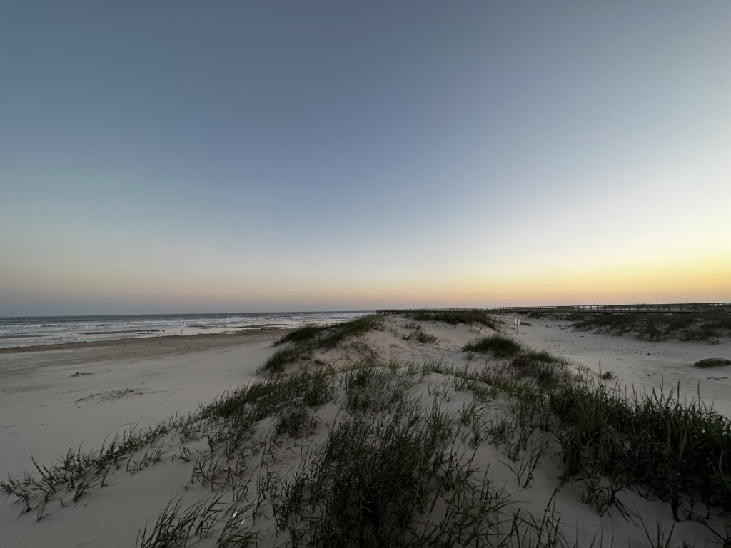 A sandy beach with grassy dunes in the foreground, gentle ocean waves in the distance, and a clear sky at sunset.