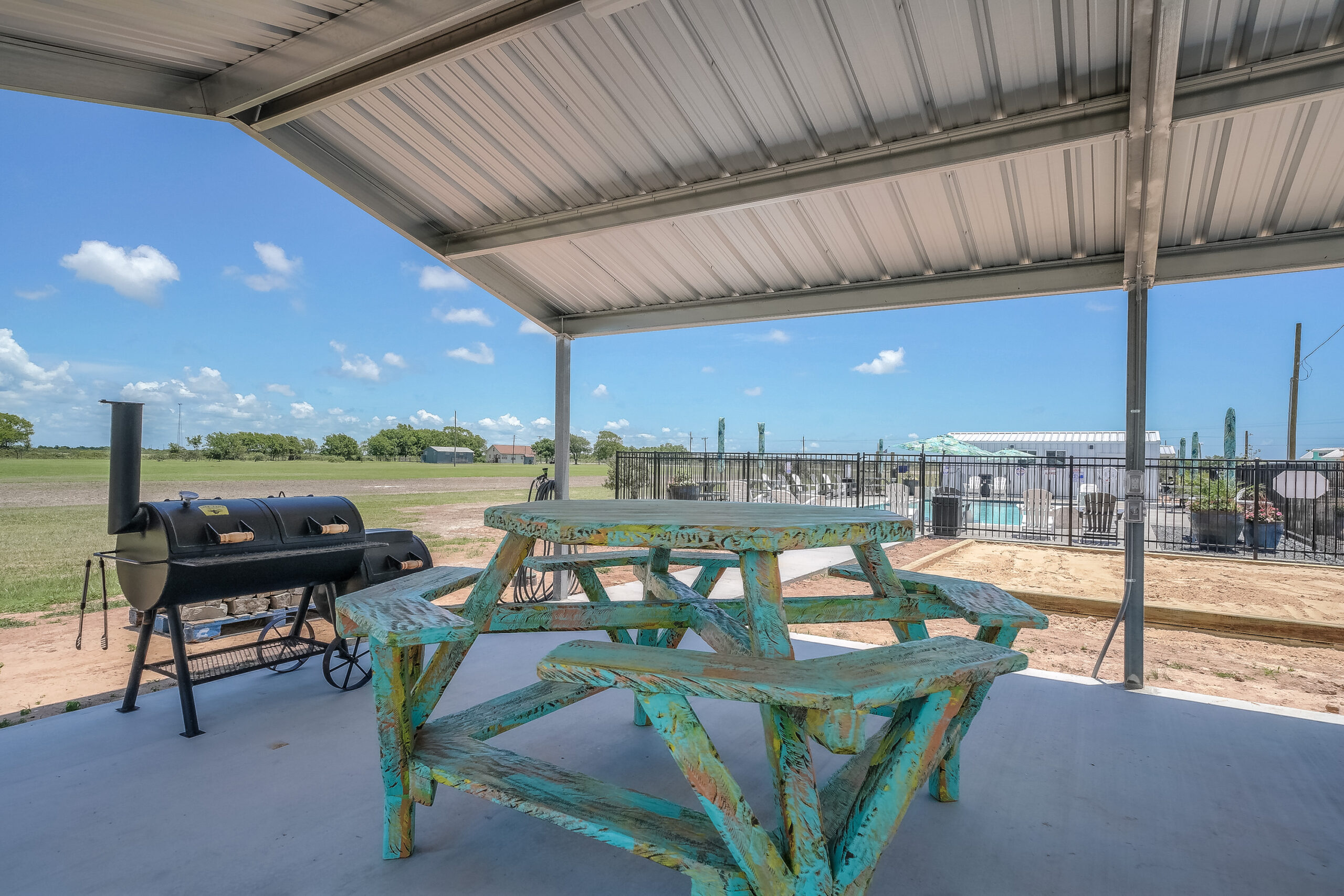 A rustic wooden picnic table and a barbecue smoker sit under a metal-roofed patio overlooking a fenced outdoor area and an open field under a blue sky.