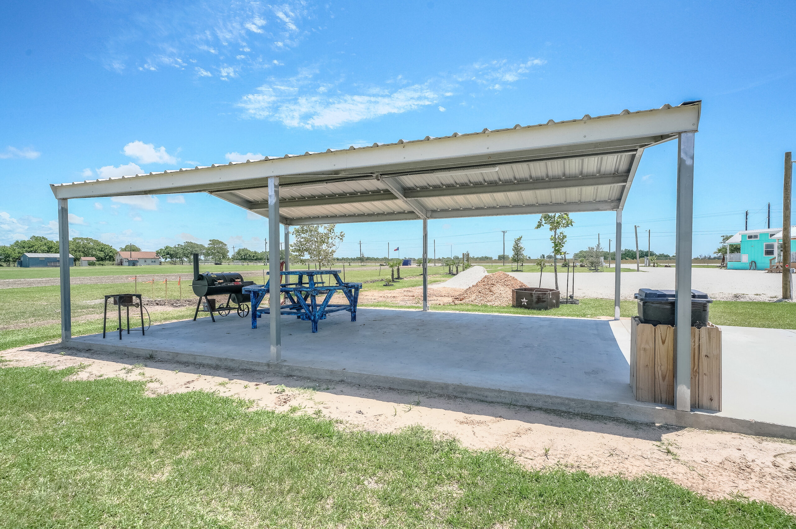 A covered outdoor pavilion with a metal roof, housing a blue picnic table and a barbecue grill, situated on a concrete slab in a grassy area under a clear sky.